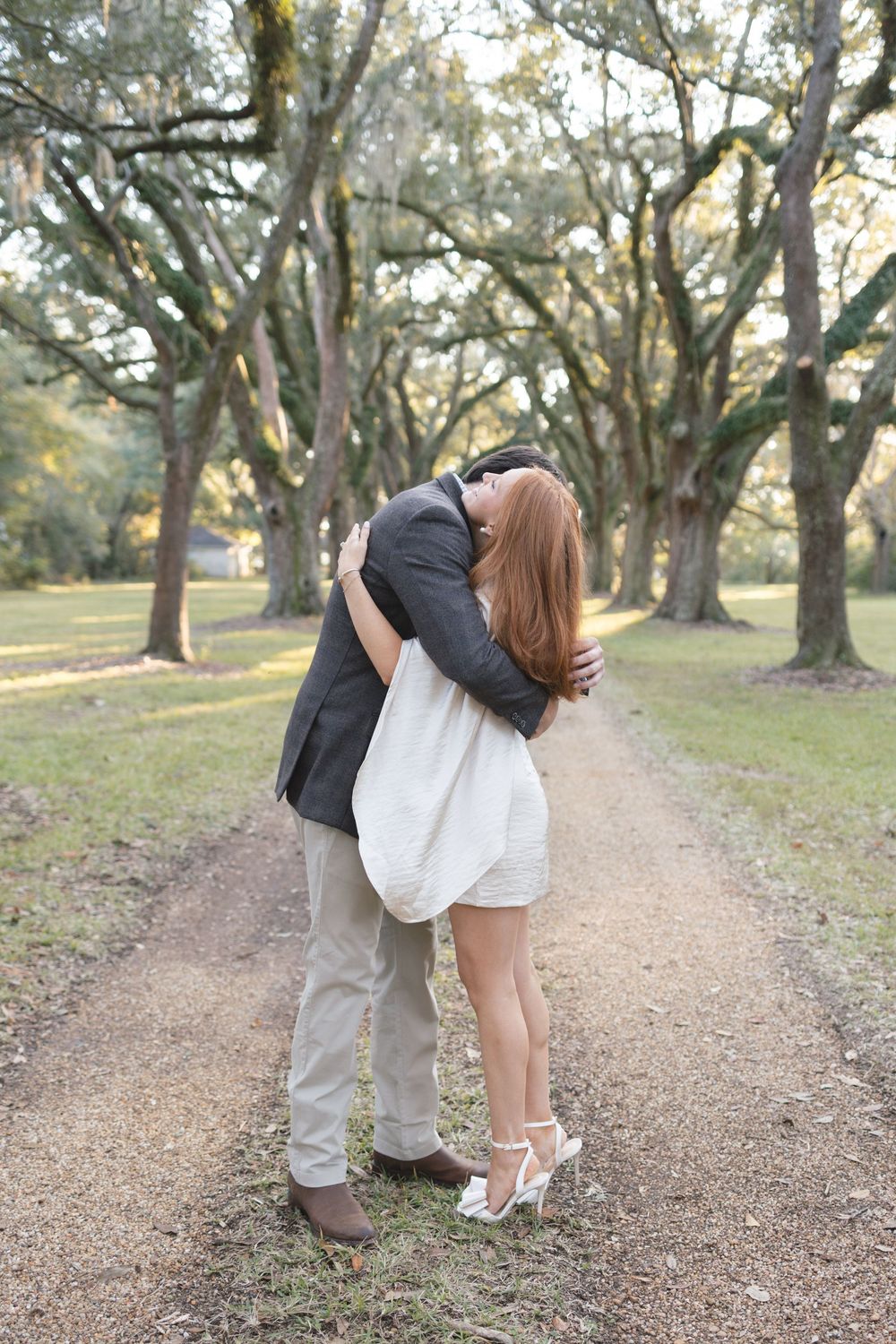 Engaged couple sharing romantic kiss on picturesque tree-lined path during outdoor photo session.