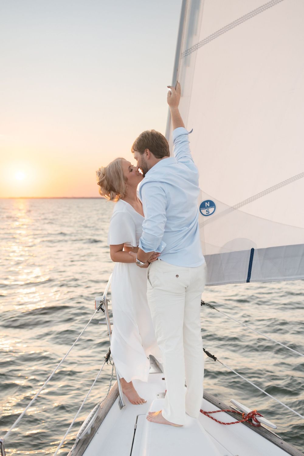 Couple kissing on sailboat bow at golden sunset with sail and ocean in background.
