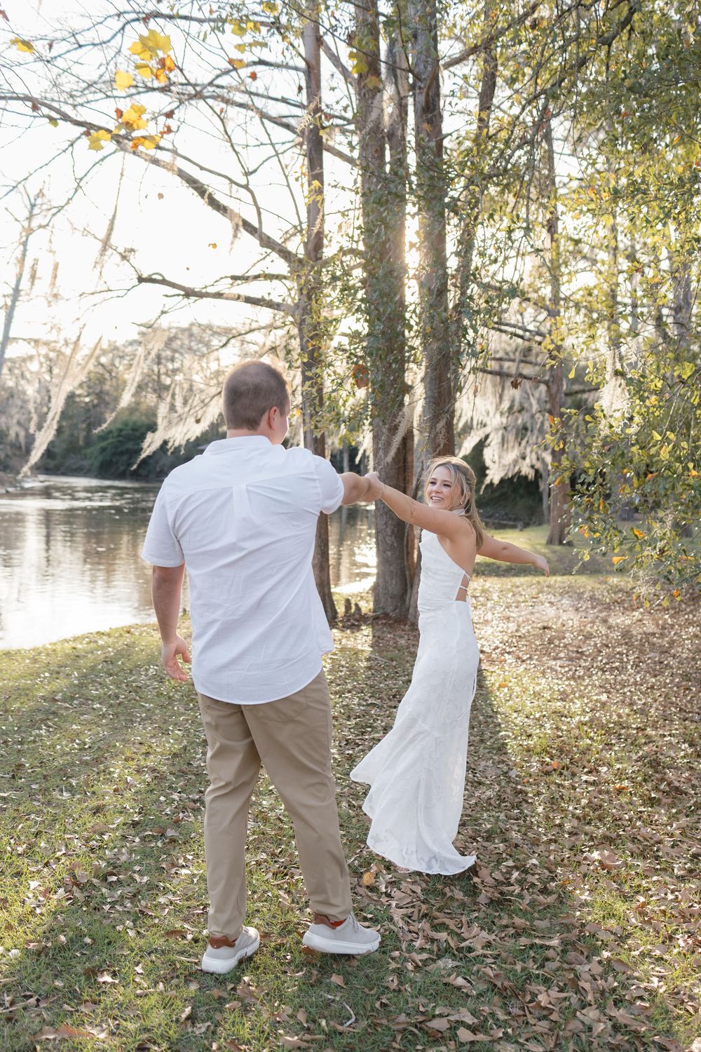 Joyful bride in white dress dancing with groom near a serene lake surrounded by autumn trees.