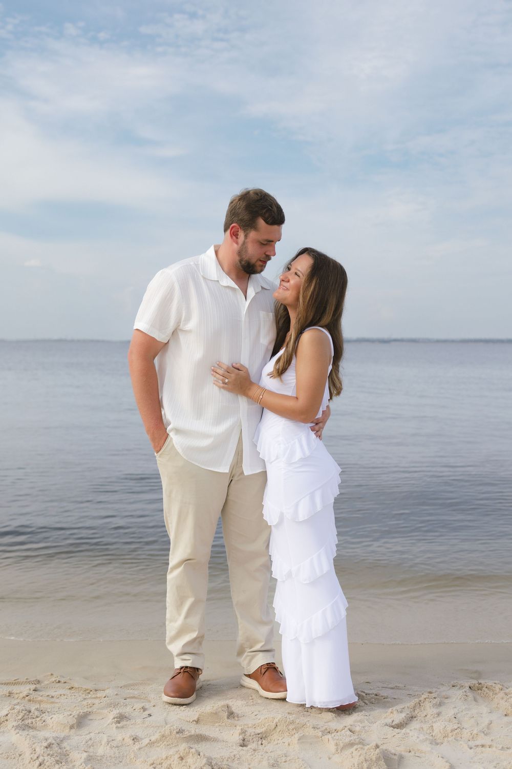 Couple in white outfits standing close together on a sandy beach with calm water behind them.