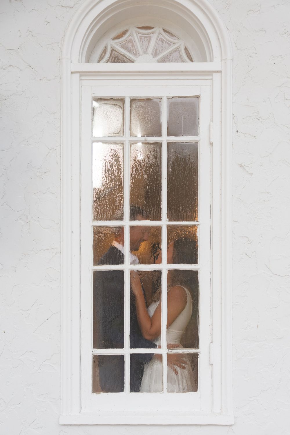 Bride and groom sharing a kiss seen through a white arched church window on their wedding day.