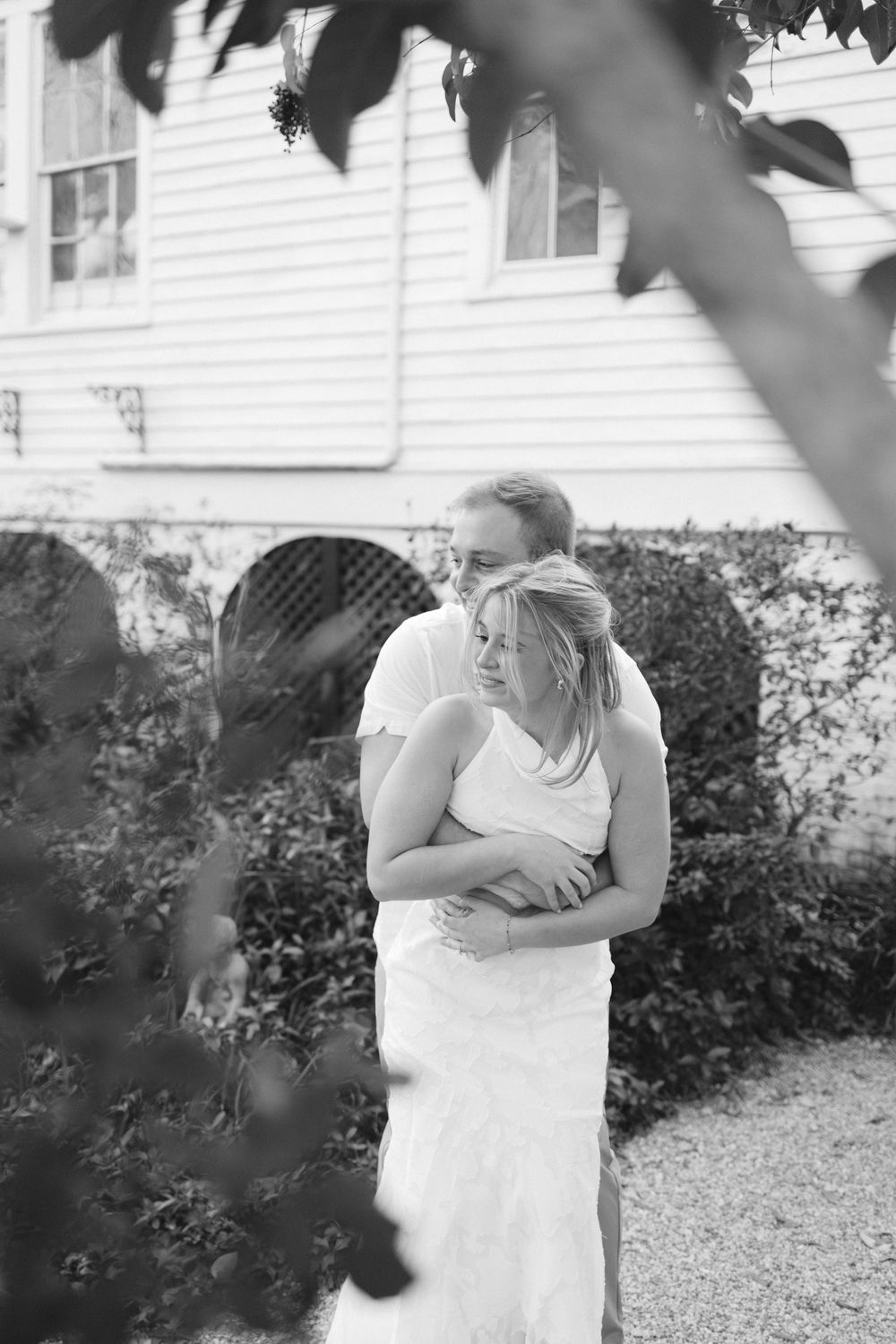 Black and white photo of couple embracing lovingly outside a white historic building with arched details.