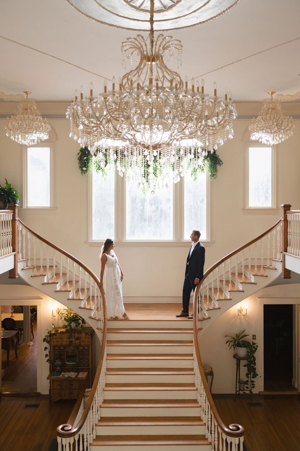 Bride and groom stand on elegant dual staircase beneath crystal chandeliers in a grand wedding venue foyer.