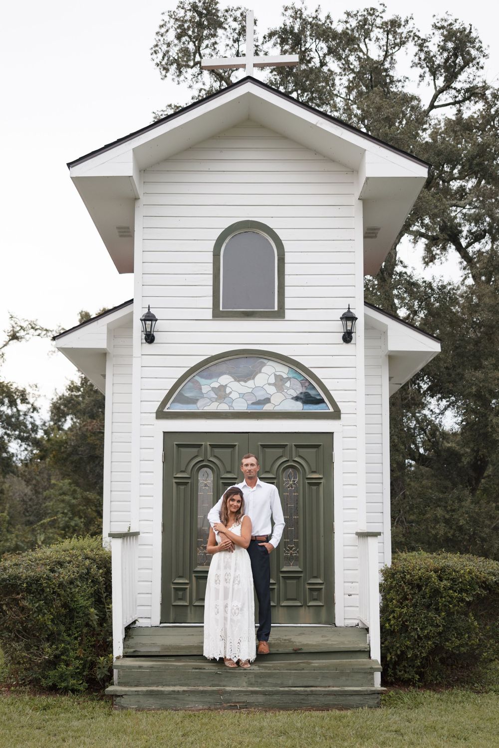Couple poses in front of a charming white chapel with green doors and stained glass window surrounded by lush trees.