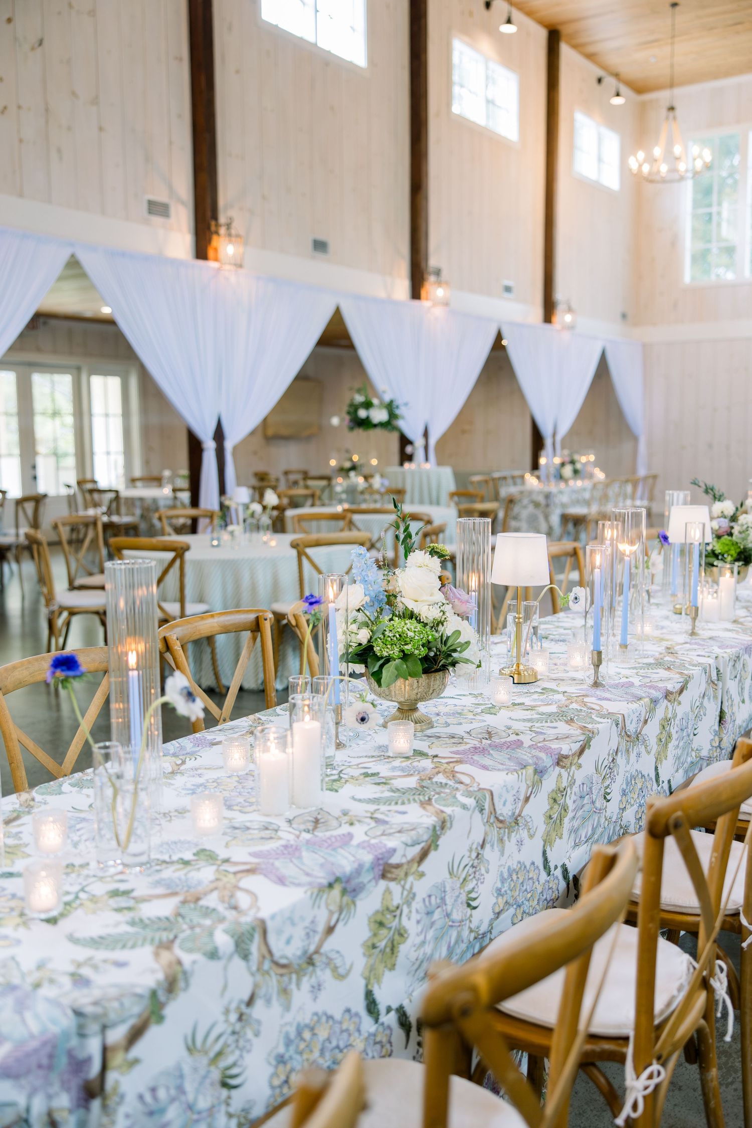 Elegant wedding reception hall with floral tablecloths, candles, wood cross-back chairs, and white draping decor.