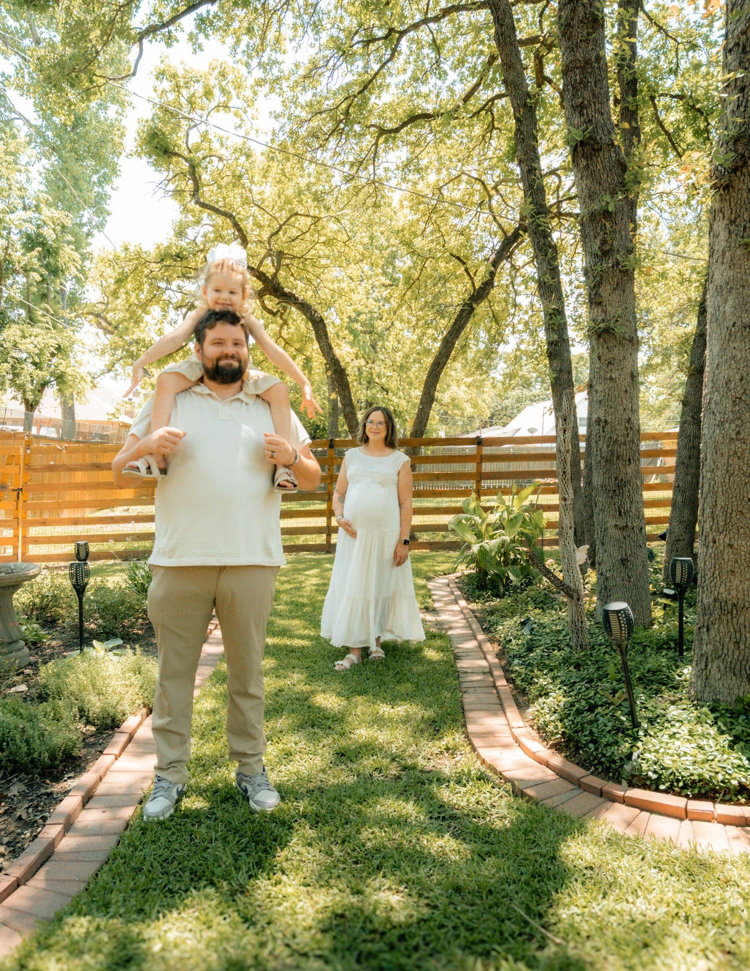 A family poses in a lush backyard garden path, father carrying toddler on shoulders, pregnant mother in white dress behind them.