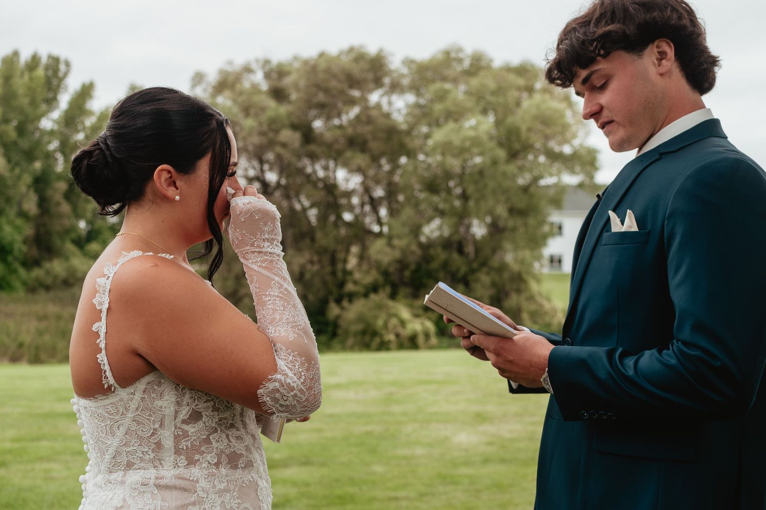 Bride wiping tears while groom reads wedding vows outdoors, wearing lace dress and teal suit.