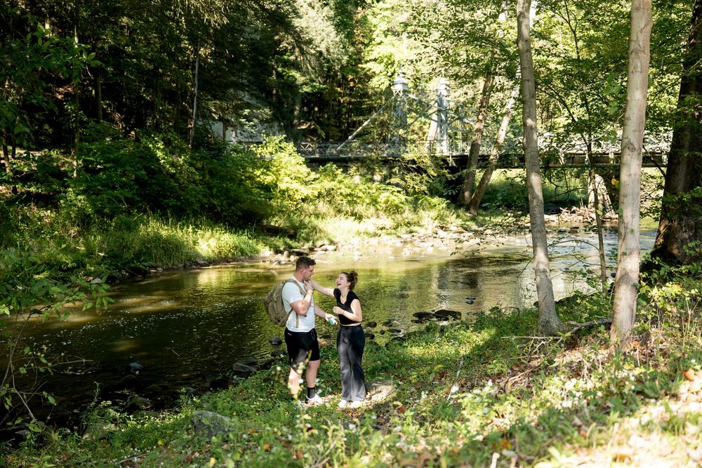 Two people stand by a peaceful forest creek with a bridge in the background on a sunny day.