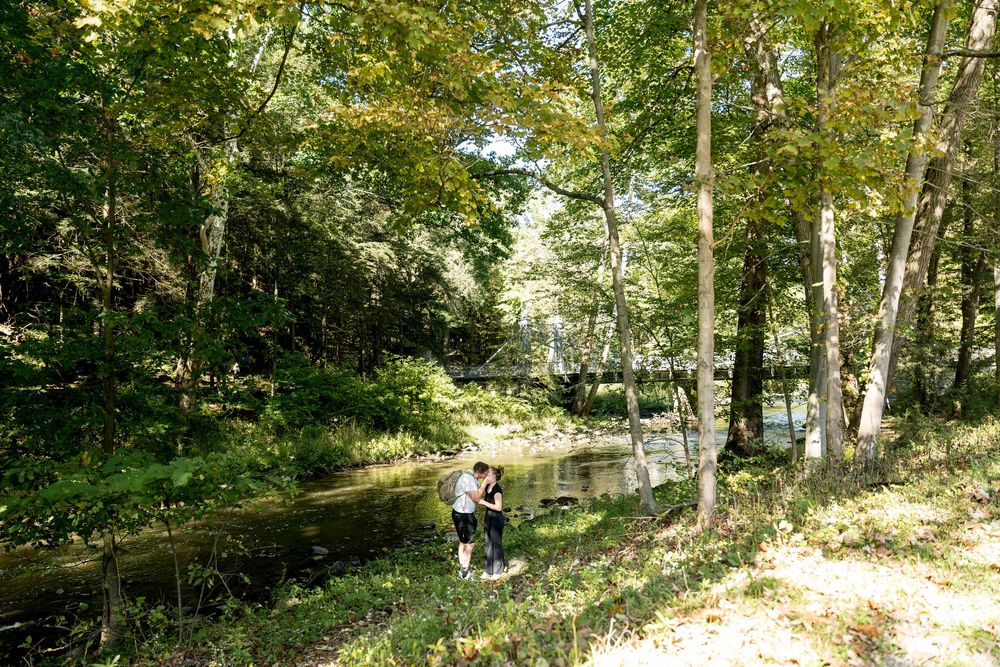 Couple embracing on a sunlit path beside a peaceful forest stream surrounded by lush green trees.