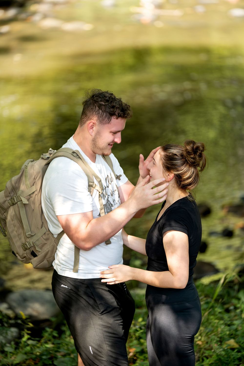 Happy couple embracing and smiling at each other beside a sunlit forest creek after proposal.