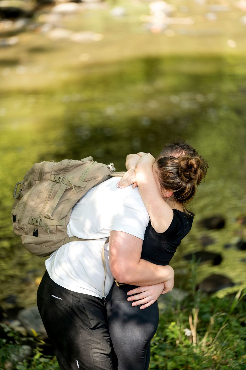 Couple sharing a heartfelt embrace near a scenic rocky river in a sunlit forest.