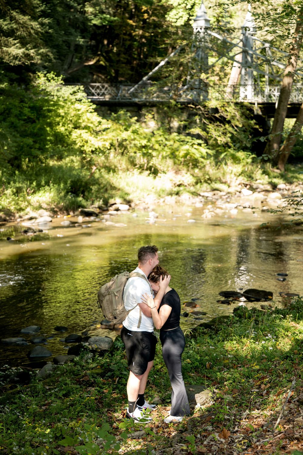 Couple sharing a romantic kiss by a peaceful forest river with a bridge and lush green trees in the background.