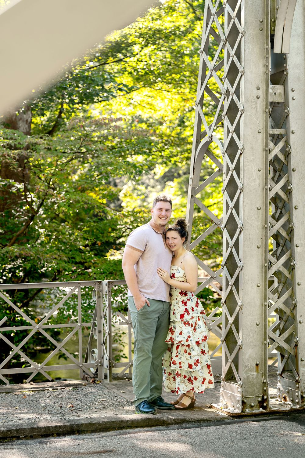 Couple embracing on a white metal bridge surrounded by lush green trees in a sunny park.