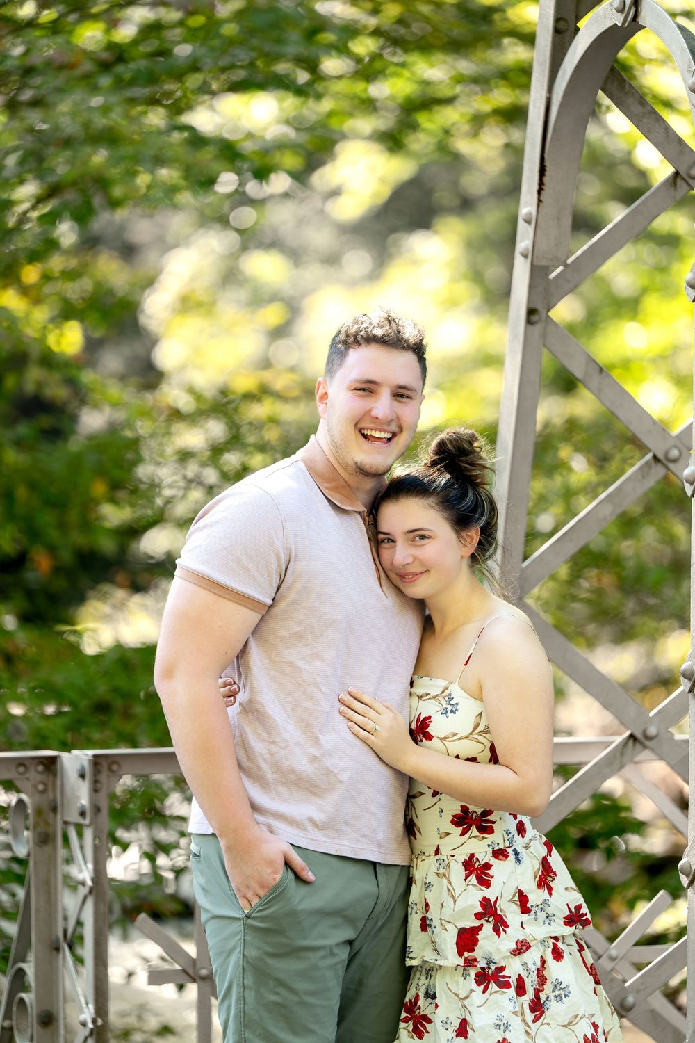 Smiling couple posing together on a metal bridge surrounded by green trees in summer.