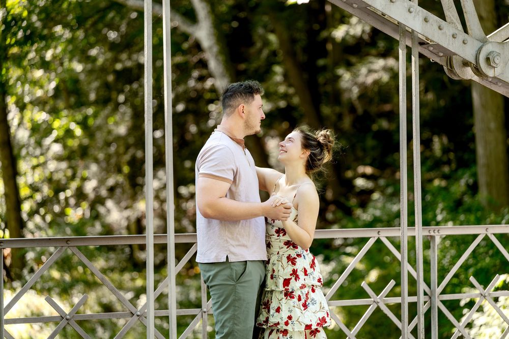 A romantic couple shares a tender moment on a white metal bridge surrounded by lush green trees.