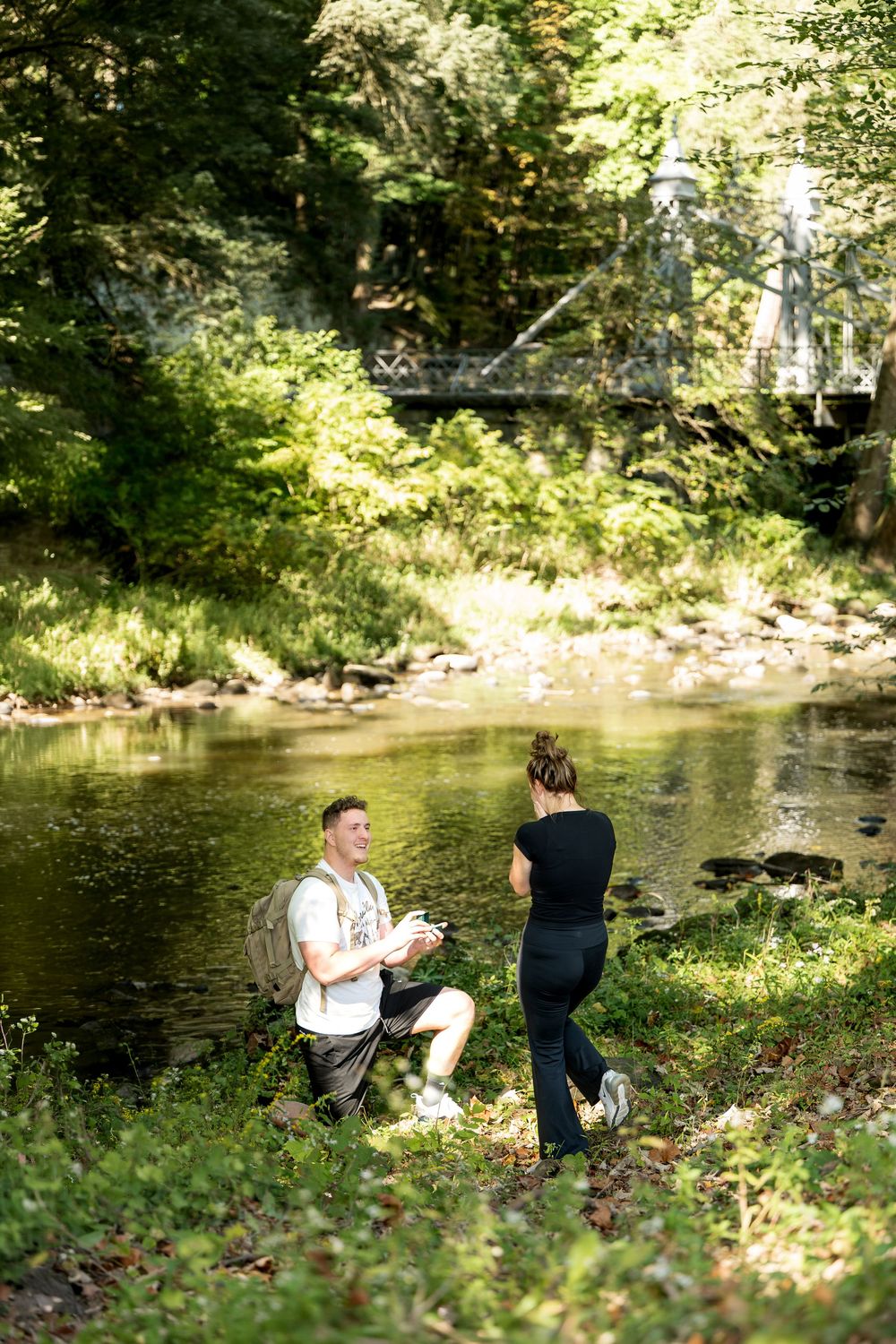 Couple sharing a romantic proposal moment beside a serene forest creek with a bridge backdrop.