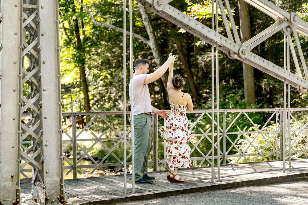 Couple dancing on a white iron bridge surrounded by lush green trees in a romantic outdoor setting.