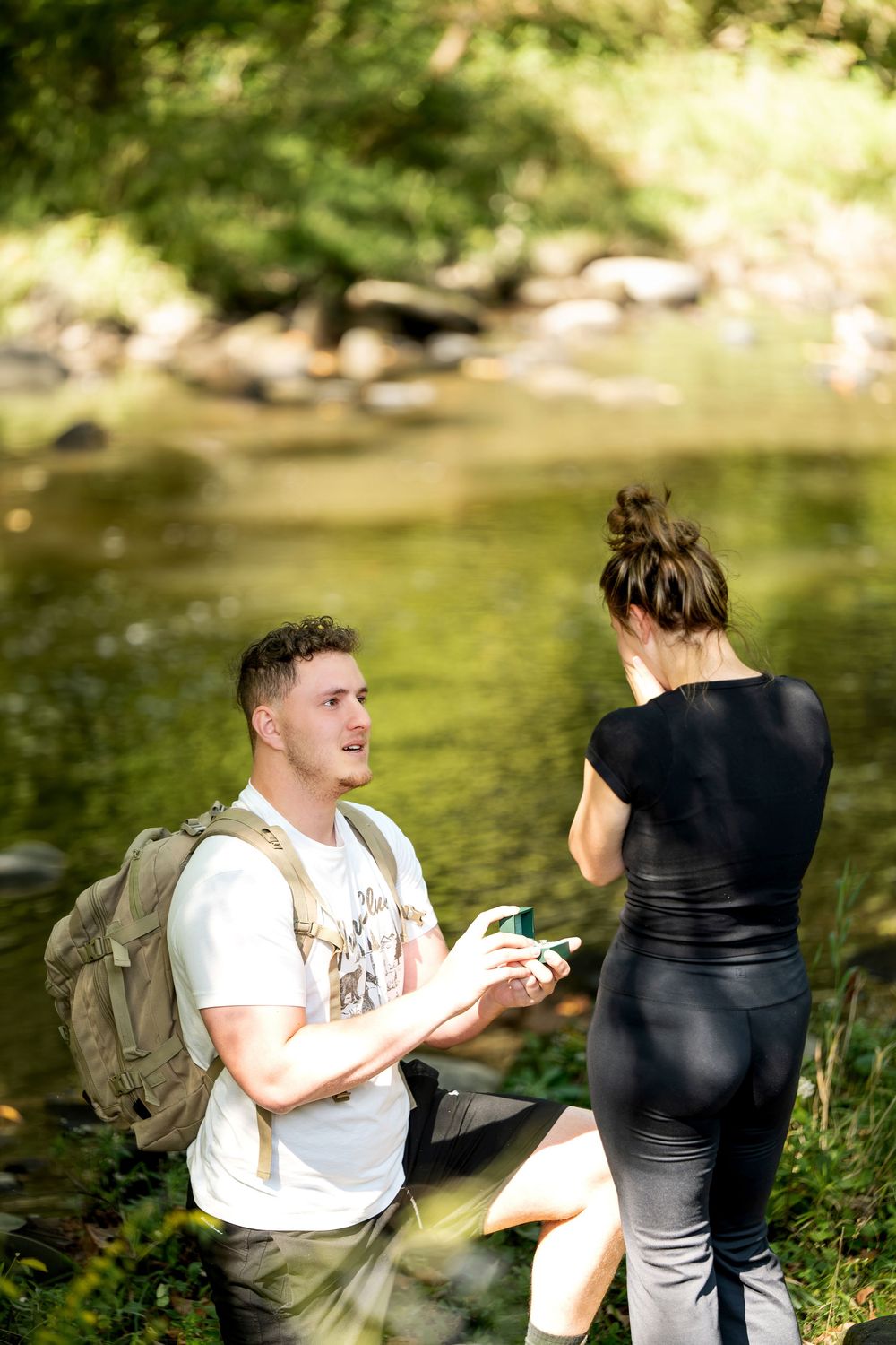 Man on one knee proposing to woman by a rocky creek in a lush green forest setting.