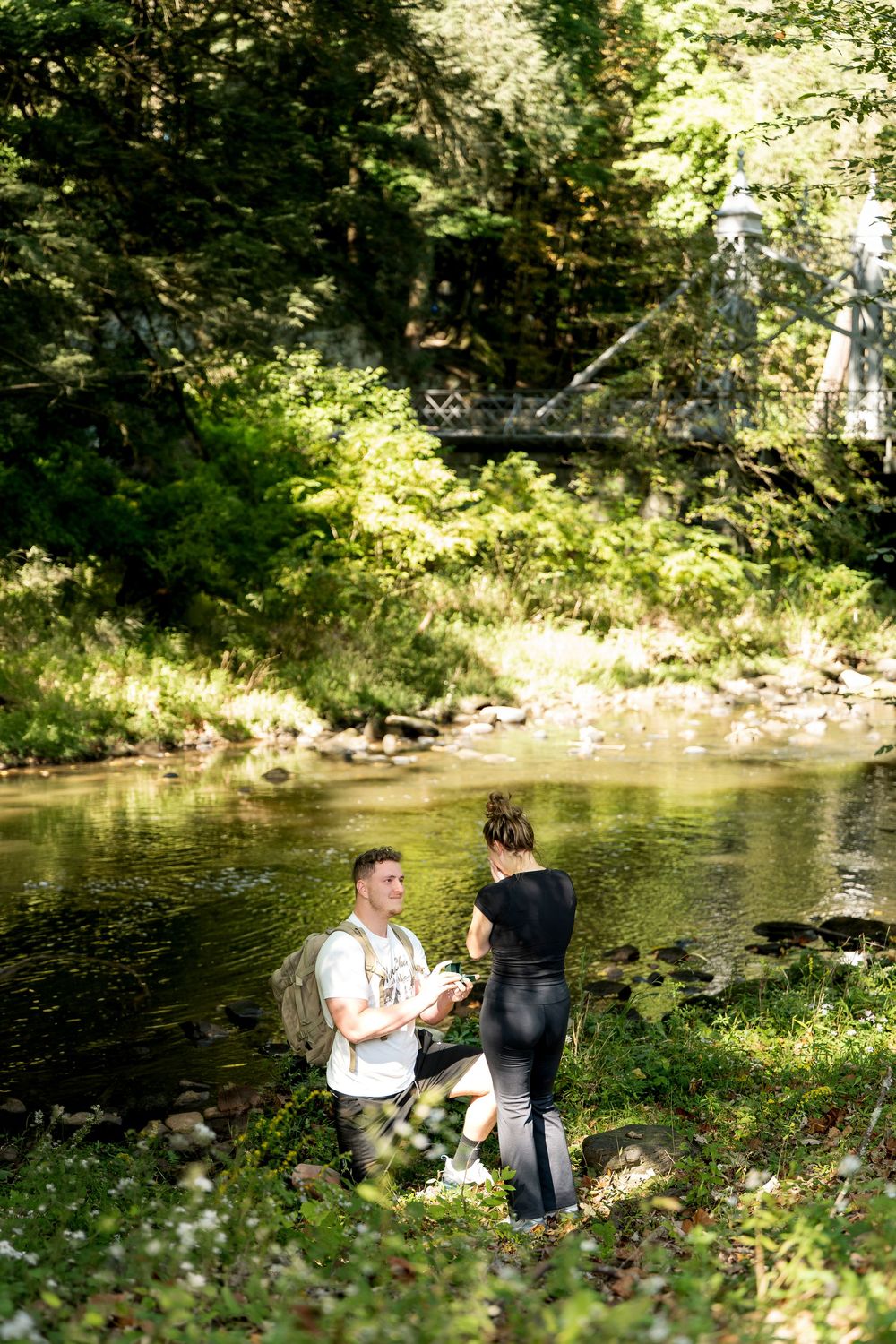 Man kneeling by a forest creek proposing to his partner during a scenic outdoor hike.