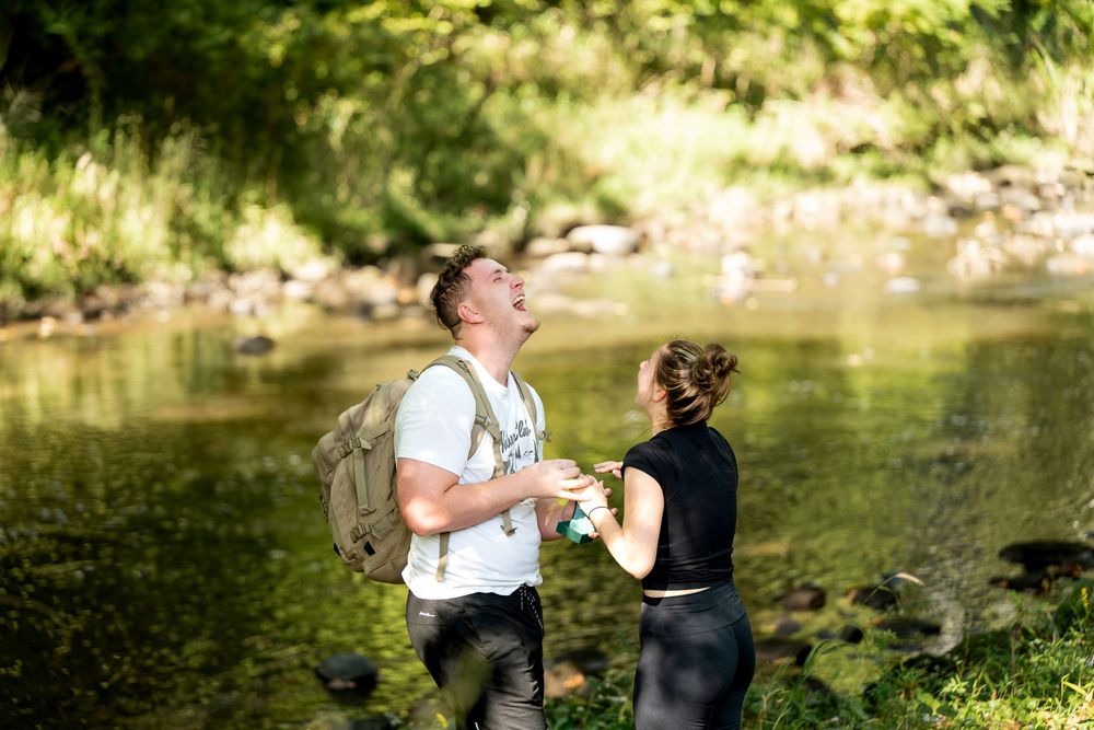 A laughing couple with a backpack enjoys a sunny hike by a rocky stream surrounded by lush green trees.