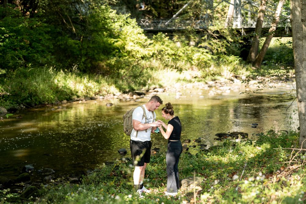 Two hikers pause by a sunlit creek surrounded by lush greenery, with a bridge visible in the background.