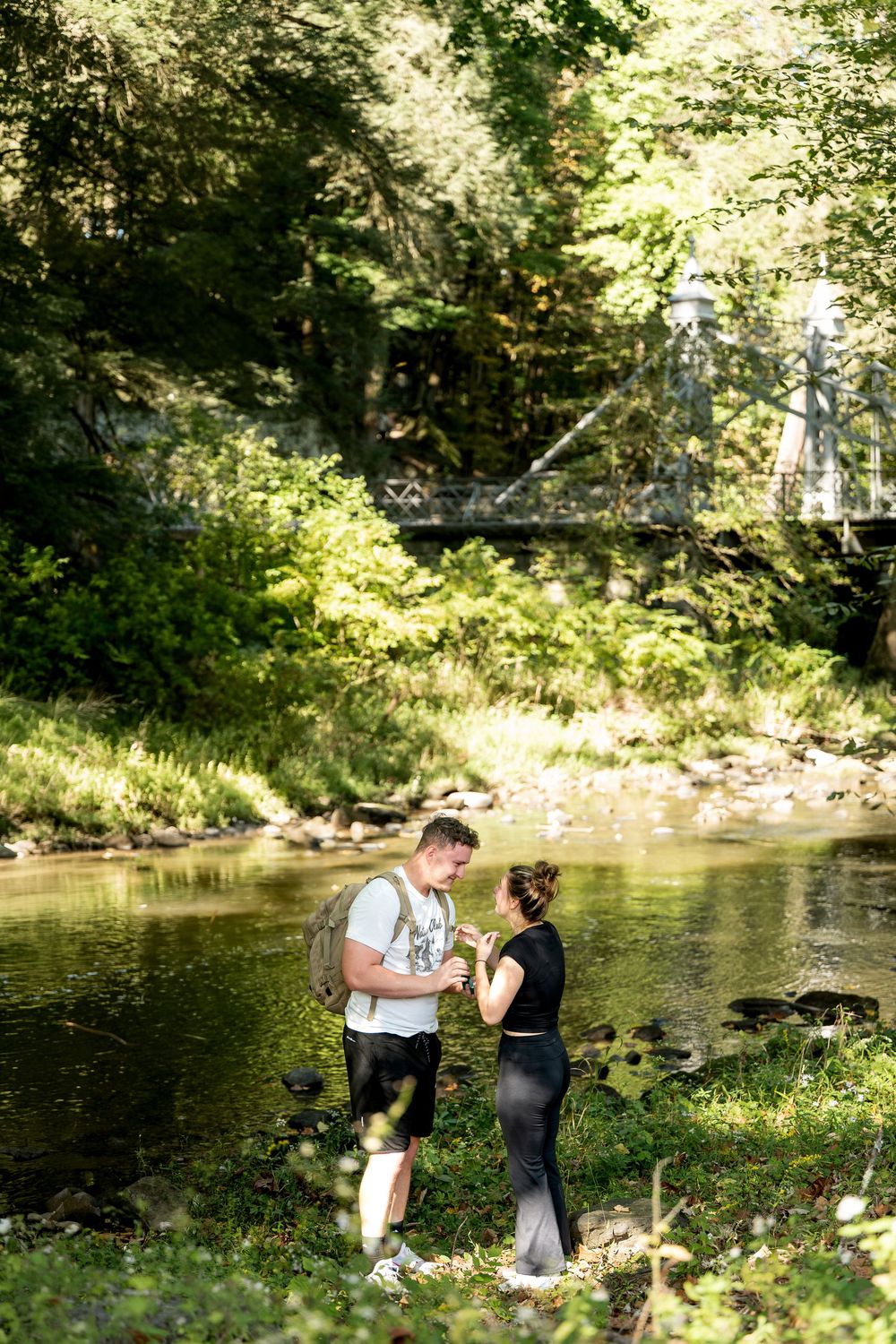 Couple standing together by a peaceful forest river, sharing an intimate engagement moment.