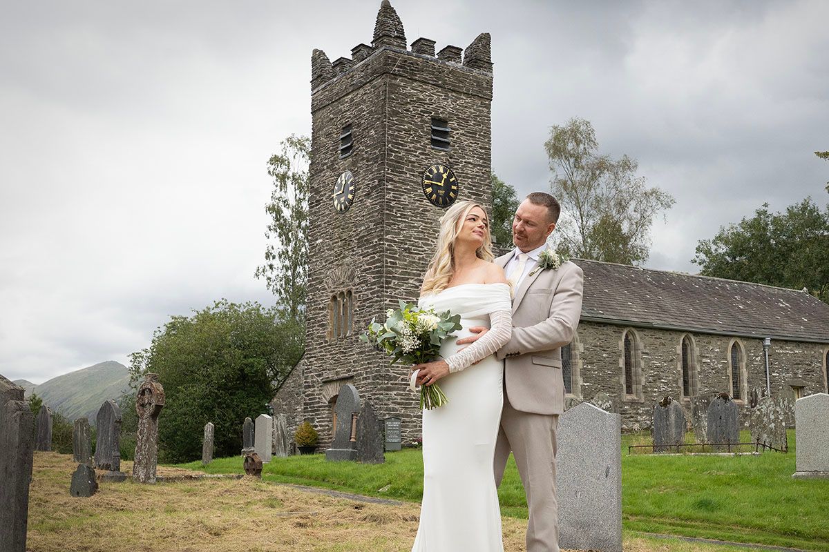 Bride and groom embracing in front of a historic stone church with clock tower and cemetery on their wedding day.