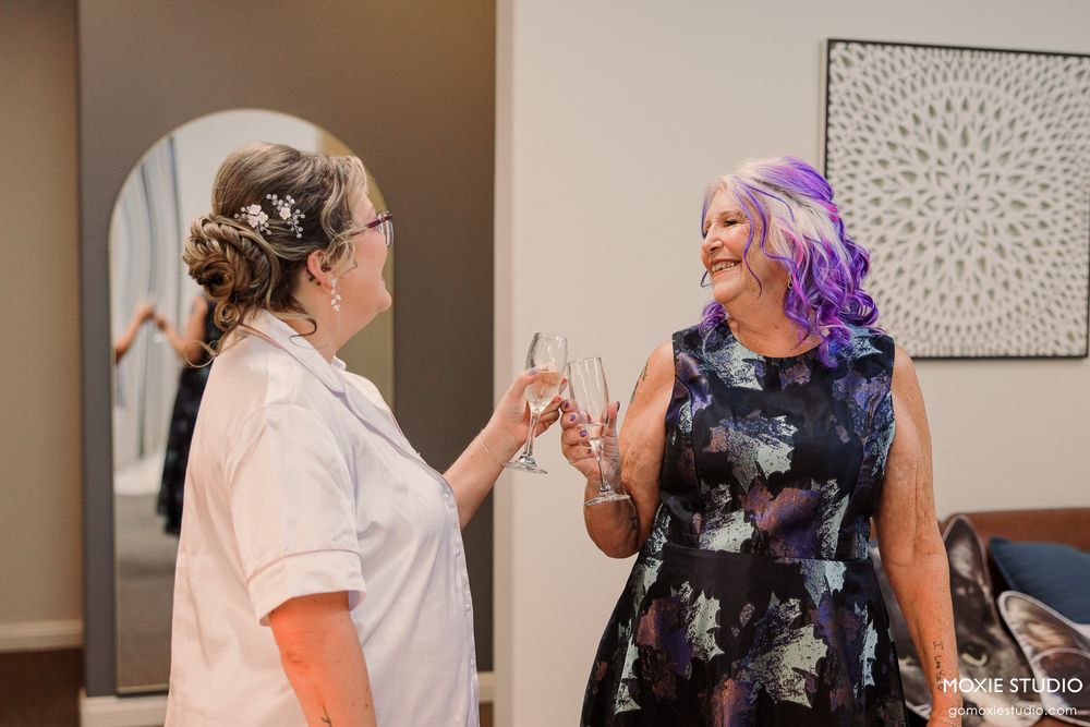 Two women toasting with champagne glasses, one in white top and one in floral dress with purple hair, celebrating together.