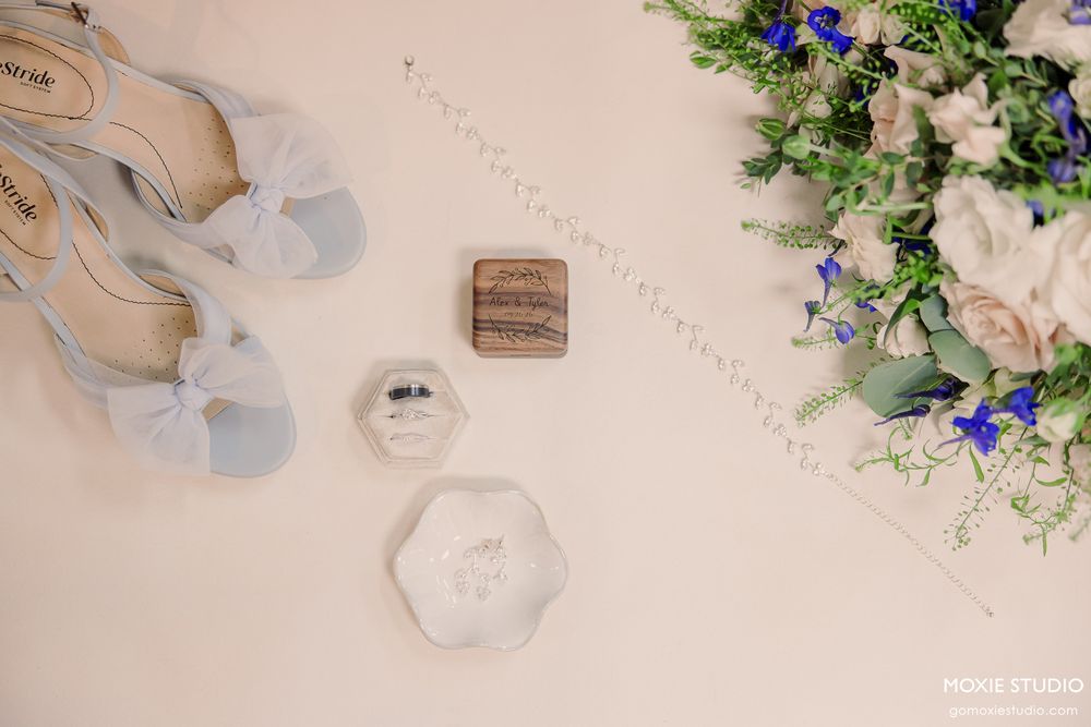 Elegant wedding flat lay with blue heels, floral bouquet, wooden ring box, and delicate jewelry on a white surface.