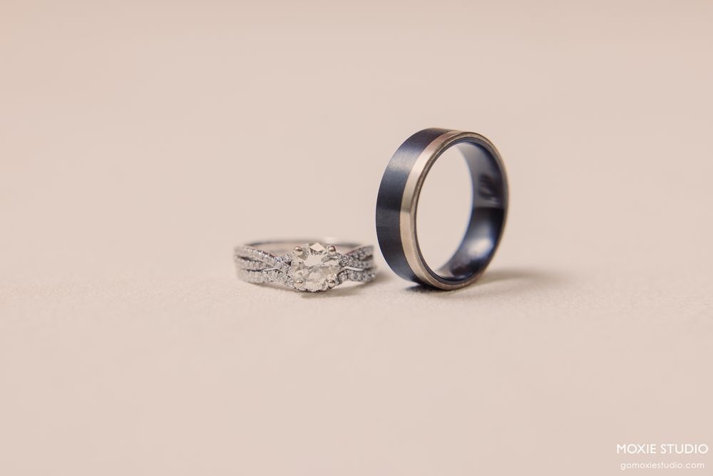 Two wedding rings displayed on a neutral background, featuring a diamond engagement ring and a dark blue mens band.