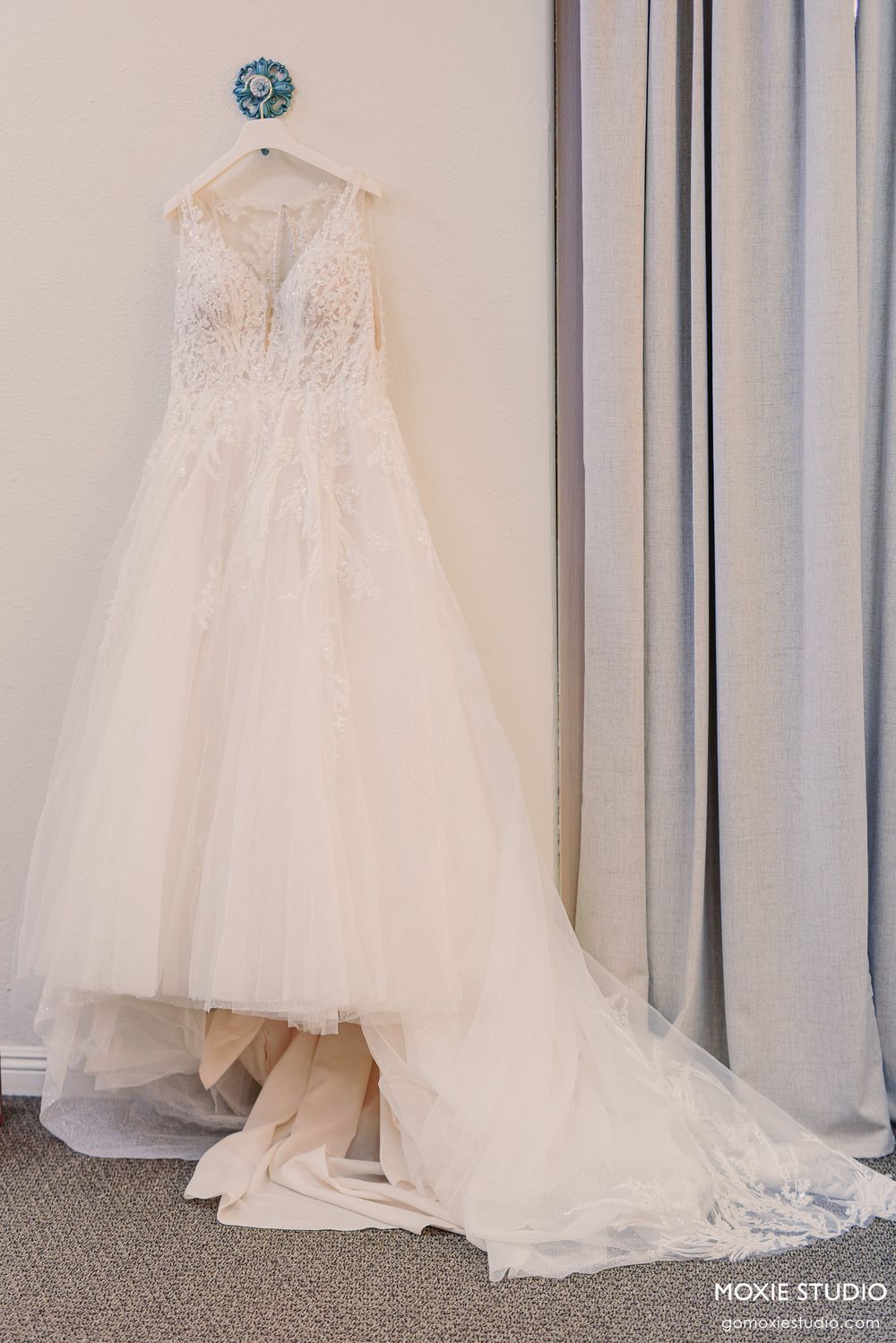 Elegant ivory lace and tulle A-line wedding dress hanging on a decorative hanger near a window with gray curtains.