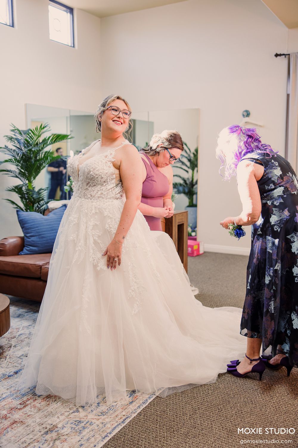 Bride in white lace ball gown smiling while attendants help with her dress in a bright dressing room.