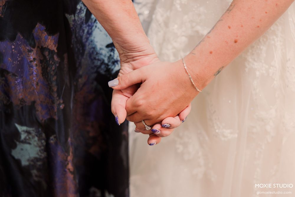 Two women holding hands at a wedding, one in a lace bridal gown and the other in a floral dress.