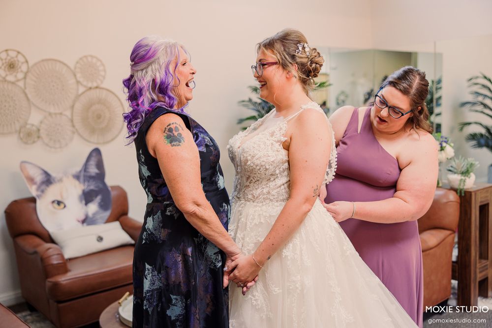 Two brides share a joyful moment holding hands in a bridal suite, one in a lace wedding gown and another in a floral dress.