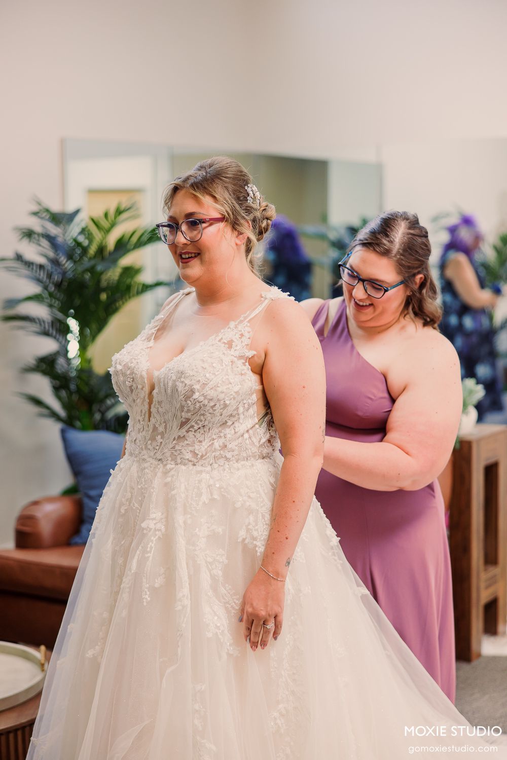 Bride smiling in lace wedding gown while bridesmaid in purple dress helps fasten the back before the ceremony.