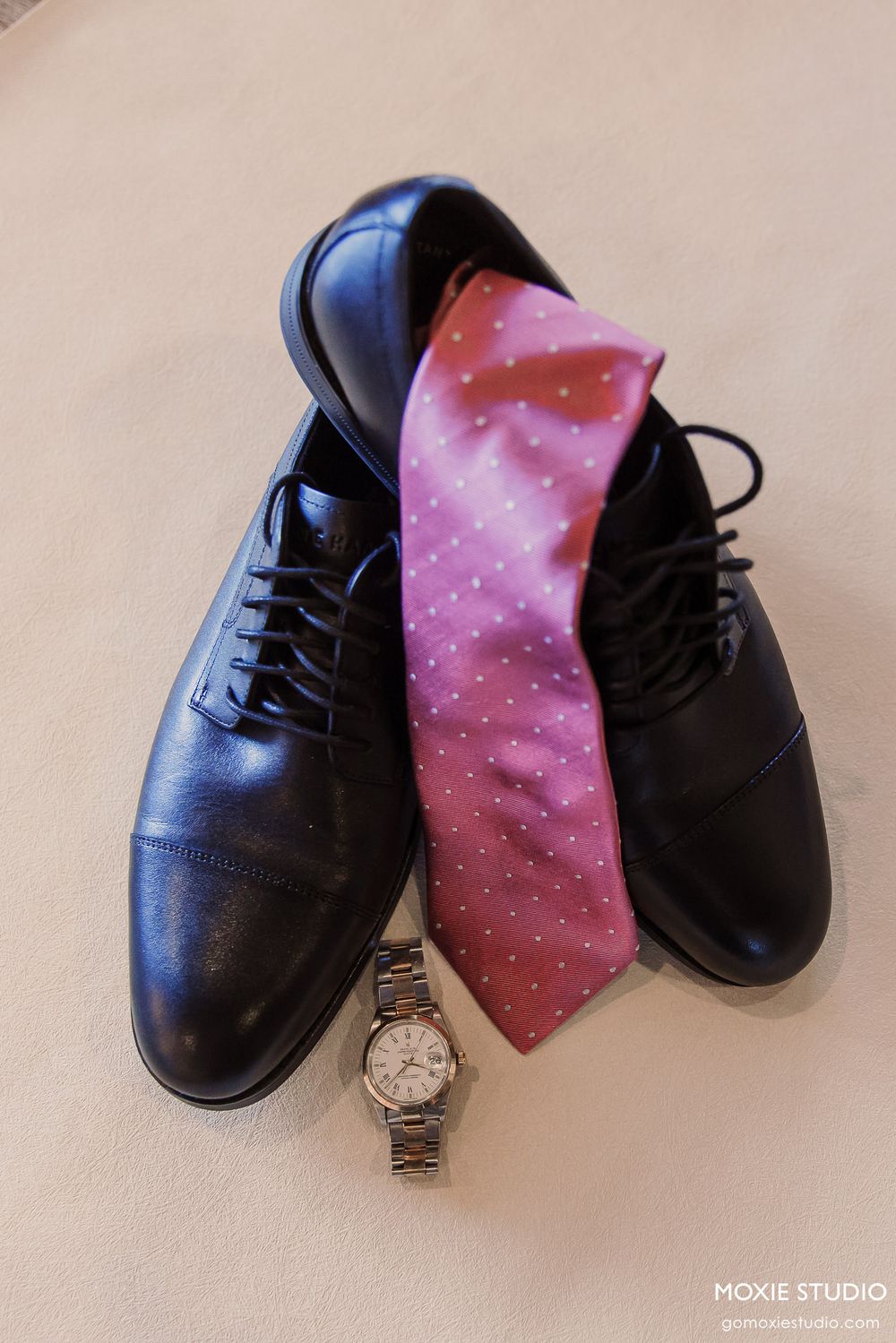 Black dress shoes with a pink polka dot tie draped over them and a silver watch placed in front on a white surface.