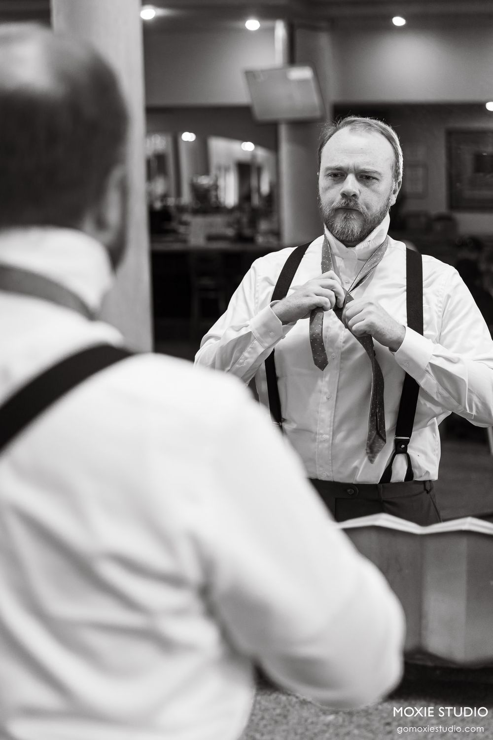 Black and white photo of a bearded groom in suspenders adjusting his tie in a mirror before his wedding.