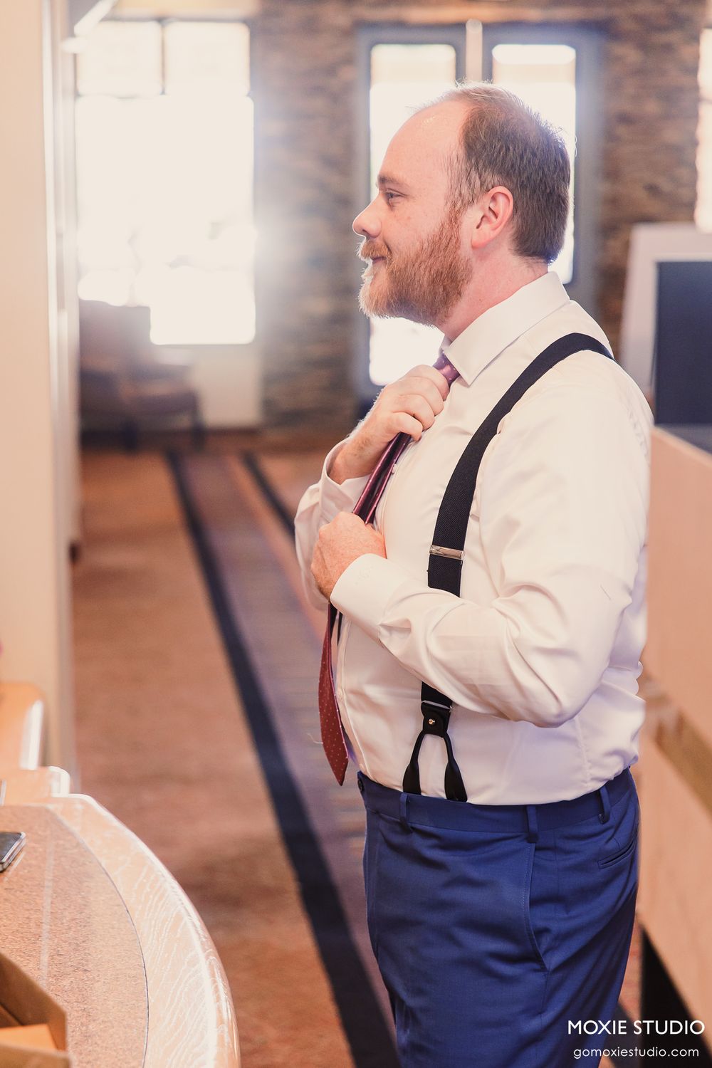 Groom adjusting his red tie wearing black suspenders and navy pants while getting ready for his wedding ceremony.