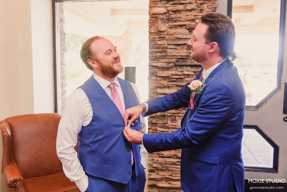 Two grooms in blue suits share a joyful moment adjusting ties before their wedding ceremony.