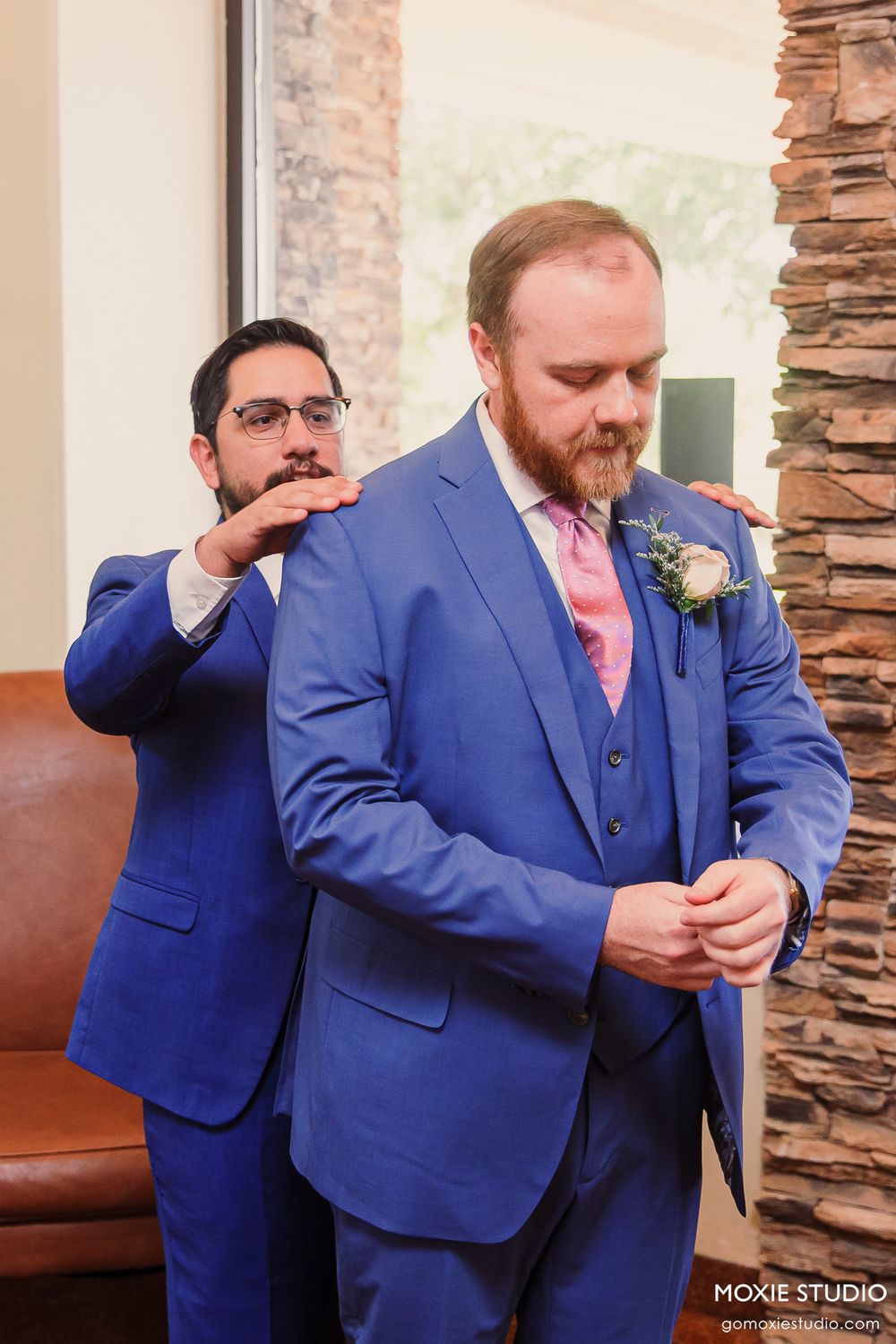 Groomsman adjusts the collar of a groom in a blue suit with pink tie and boutonniere before wedding ceremony.