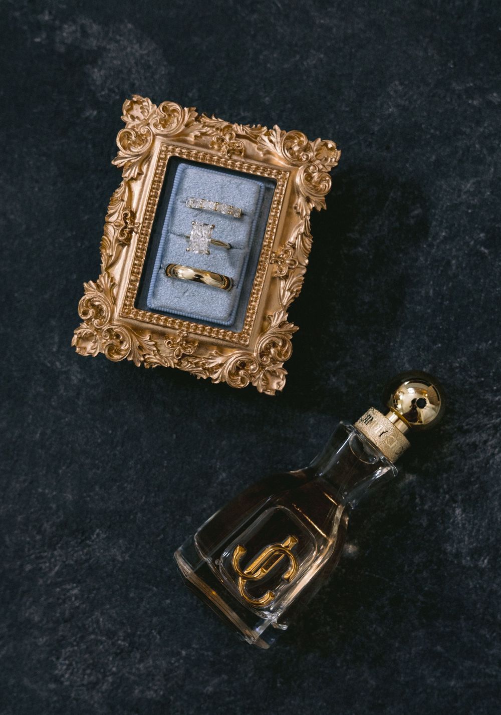 Gold ornate framed ring box displaying wedding bands and engagement ring beside a crystal perfume bottle on dark marble.