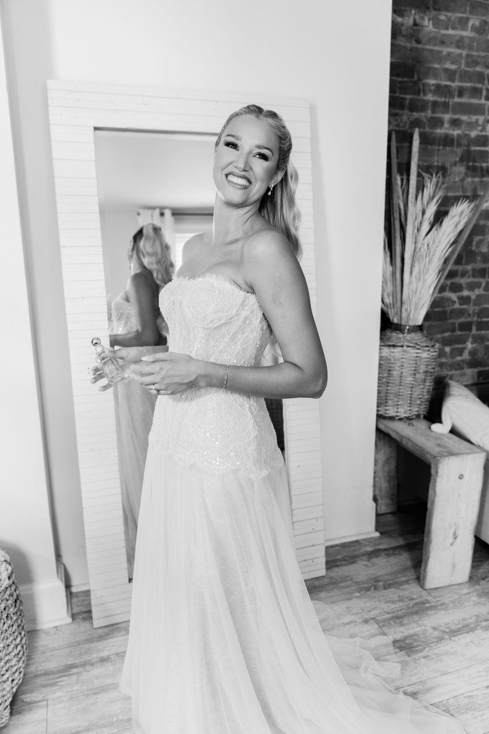 Smiling bride in strapless white wedding gown poses in front of mirror in black and white photo.