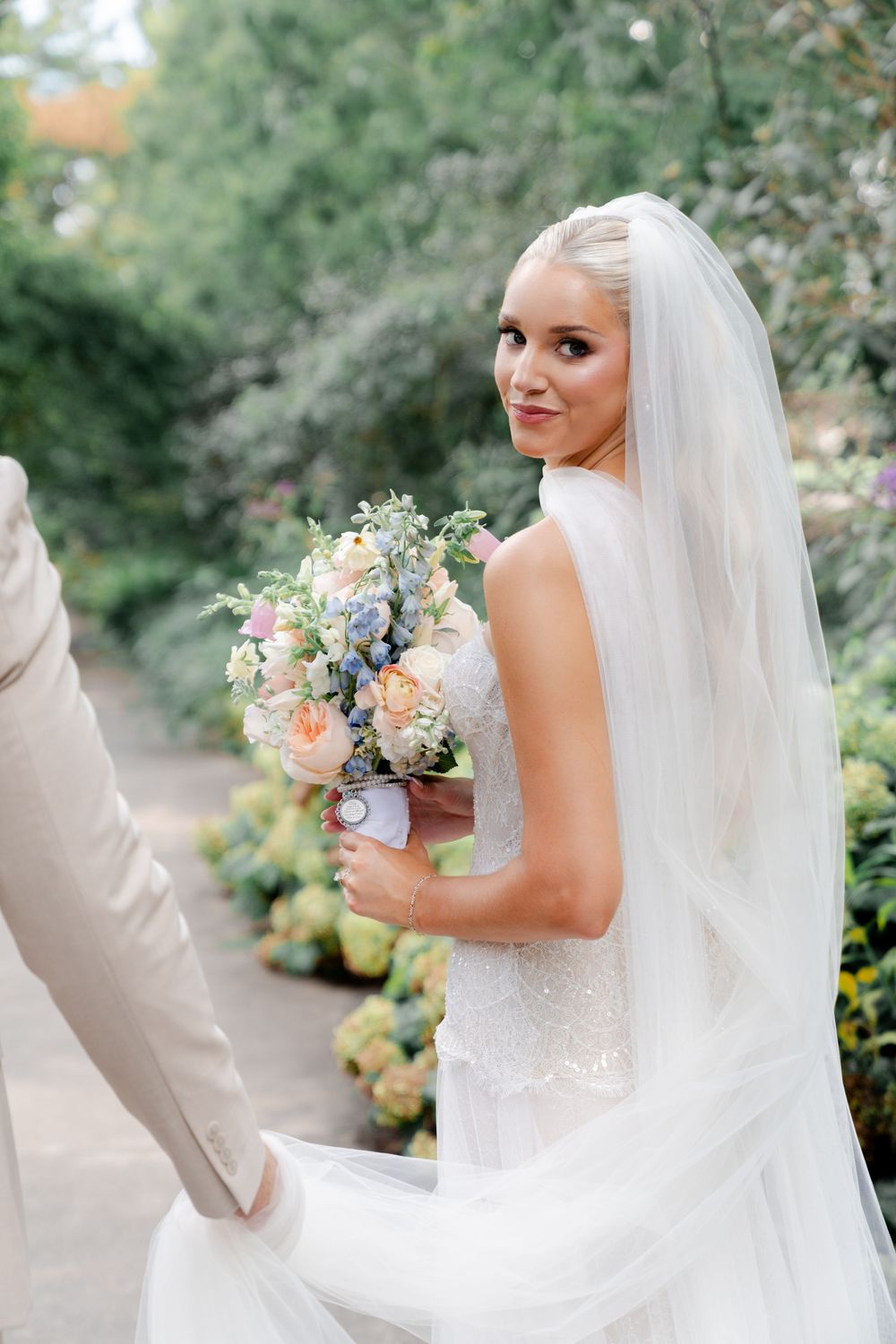 Smiling bride in white gown and veil holds pastel bouquet of peonies and hydrangeas in a lush garden setting.