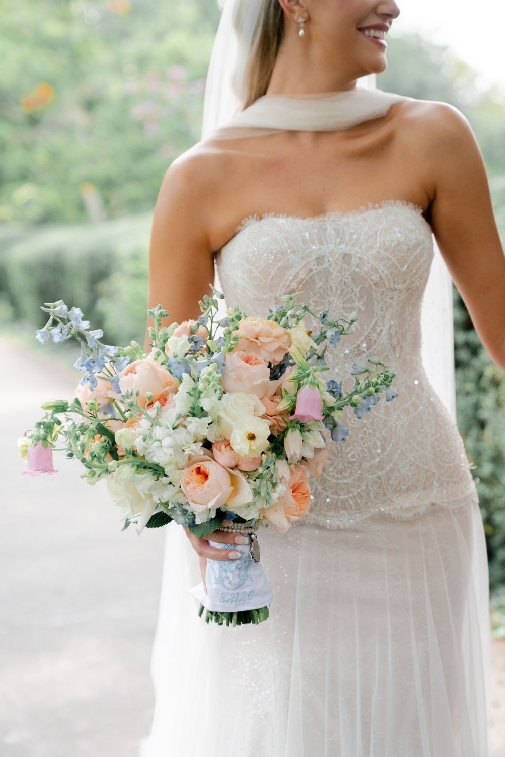Bride in strapless beaded gown holding a pastel bouquet of peach roses and blue flowers outdoors.