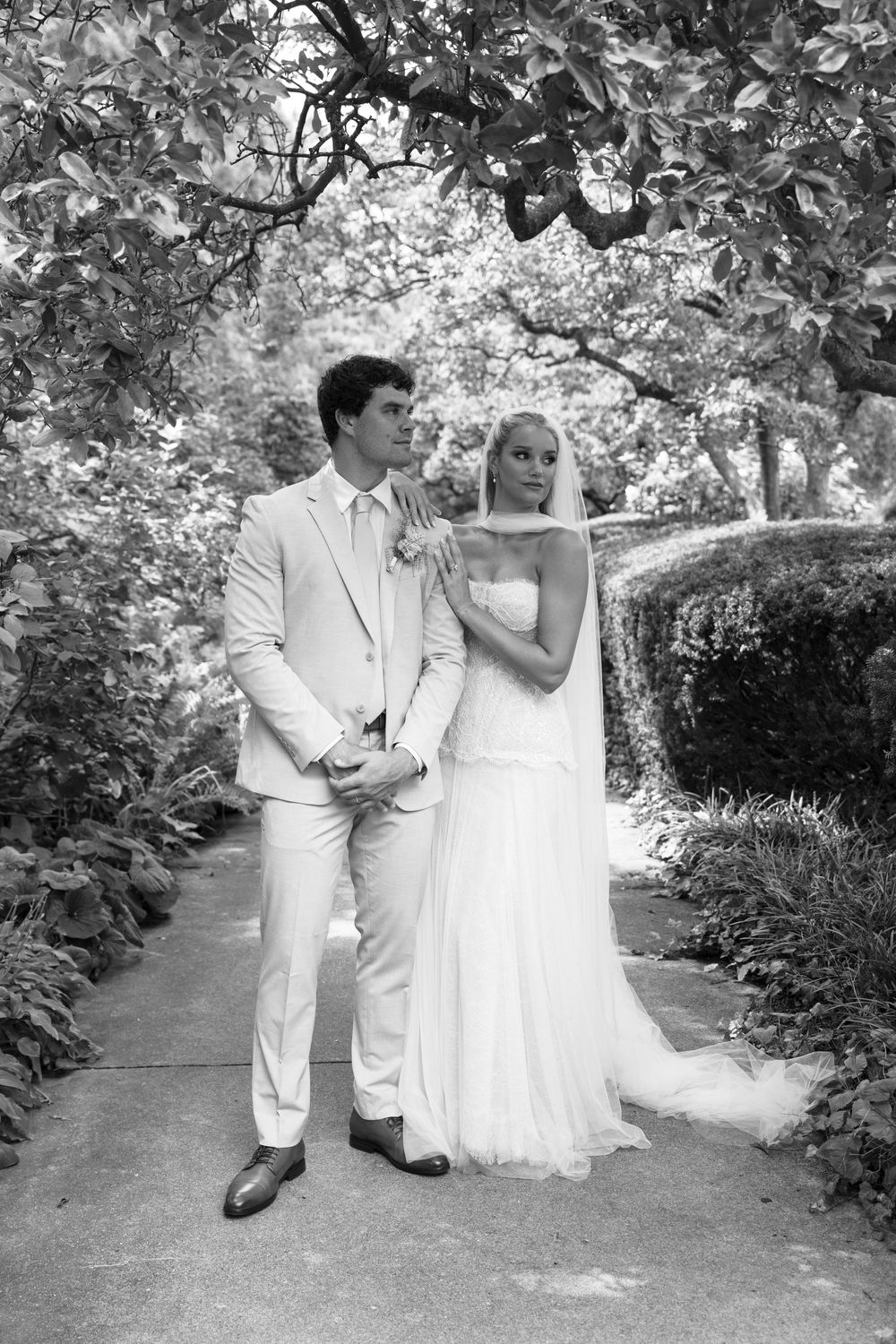 Black and white photo of a bride and groom posing in a lush garden pathway on their wedding day.