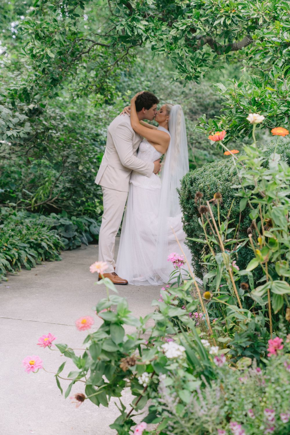 Bride and groom sharing a romantic kiss on a garden path surrounded by lush greenery and colorful blooming flowers.