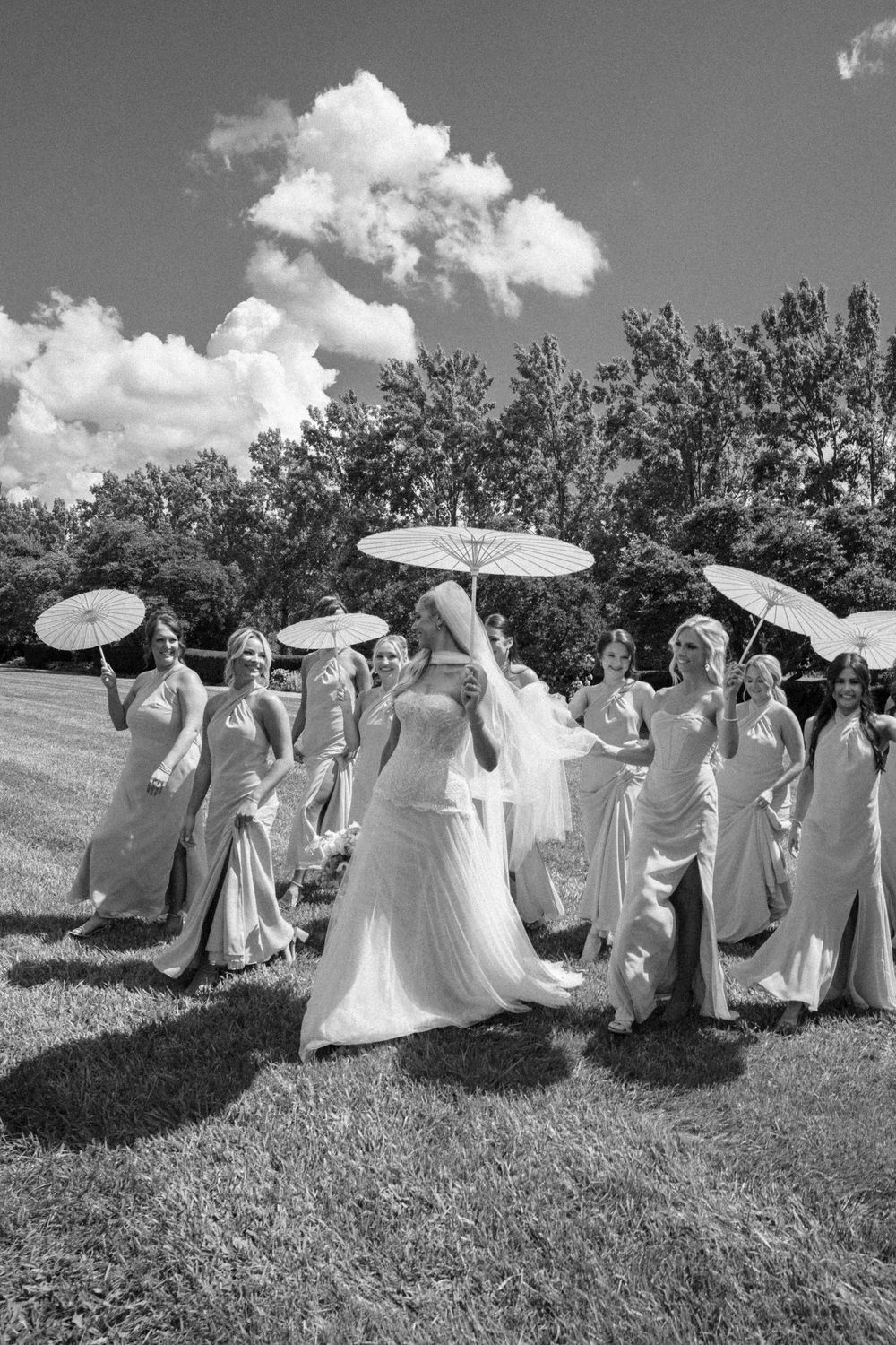 Black and white photo of a bride and bridesmaids holding parasols on a sunny day outdoors.