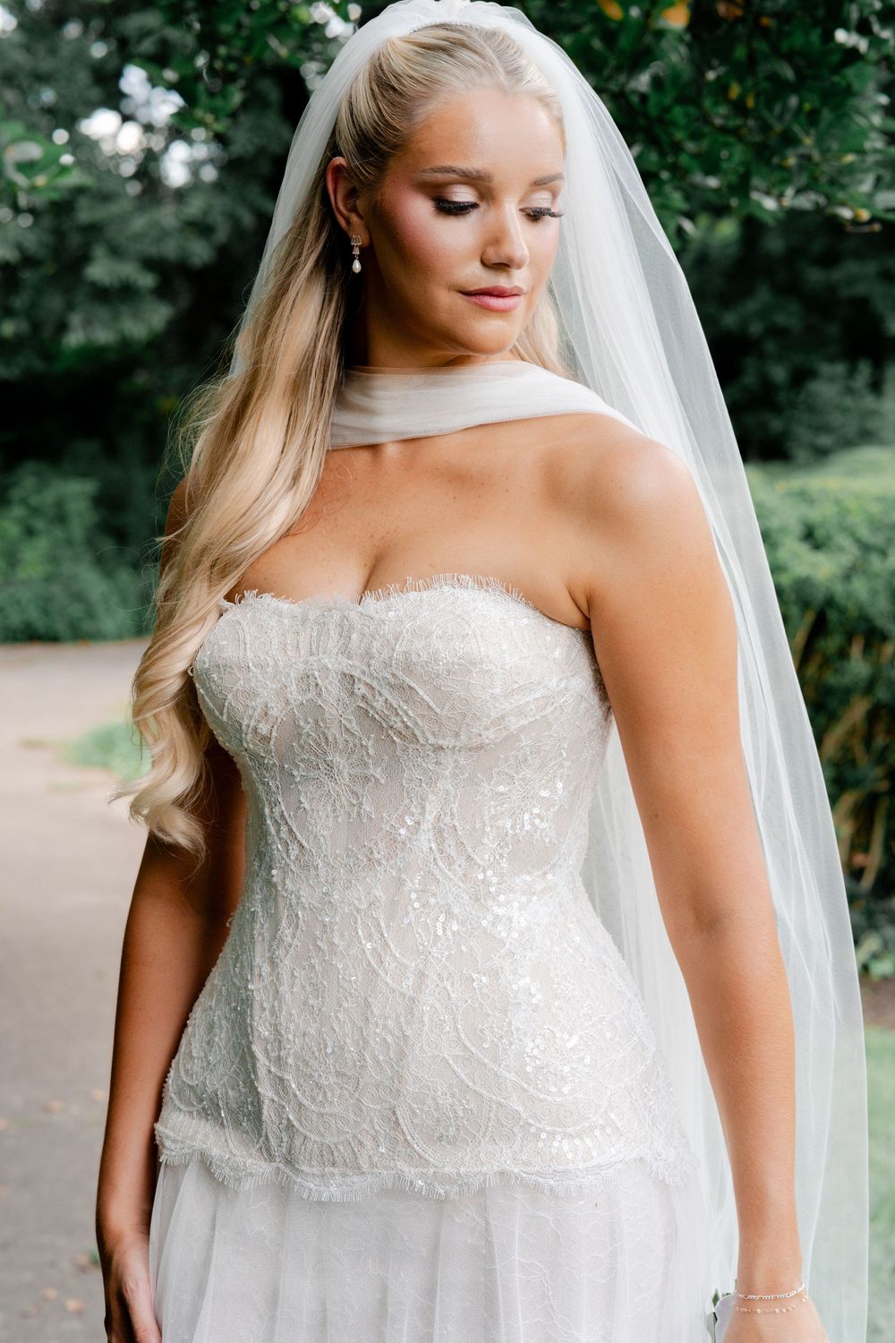 Blonde bride in a strapless lace wedding gown and veil posing outdoors in a garden.