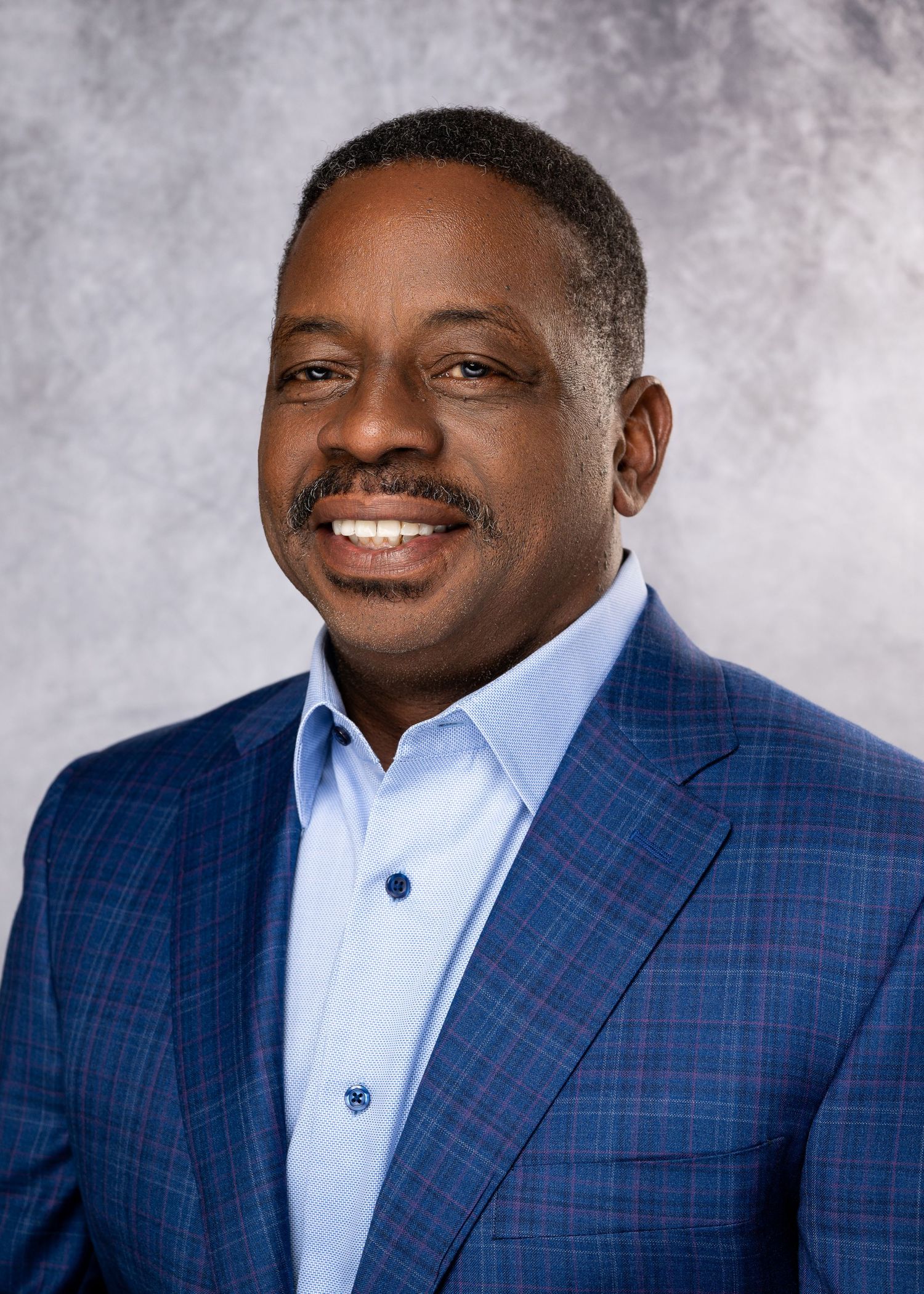 Professional headshot of a smiling man in a blue plaid blazer and light blue shirt against a gray background.