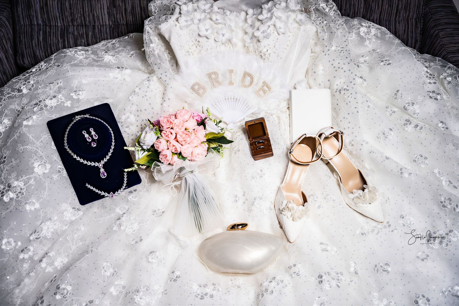 Bridal flat lay with lace wedding dress, pink bouquet, jewelry, gold rings, heels, and phone on white background.